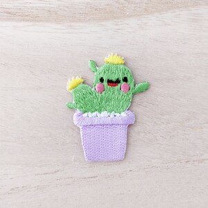 May include: Embroidered patch of a smiling green cactus with yellow accents, pink cheeks, and a black mouth. The cactus is in a lavender pot with a white trim. The patch is designed for decorative purposes.