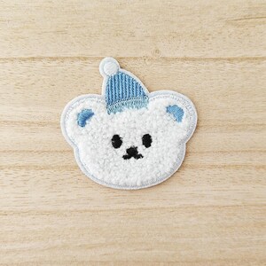 May include: Embroidered patch of a white teddy bear wearing a light blue knitted hat. The bear has black eyes and a black nose. The patch has a light blue outline and is made of a soft, textured material.