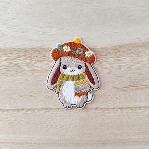 May include: Embroidered patch of a bunny wearing a brown hat decorated with flowers and a yellow pom-pom. The bunny has a colourful scarf. The patch is on a light wood grain surface. The bunny is in a seated position.