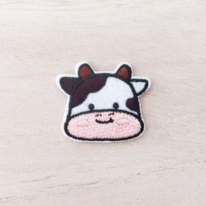 May include: A white and brown embroidered cow patch with a pink face and a happy expression.