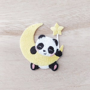 May include: A black and white panda bear holding a yellow crescent moon with a yellow star on top. The panda is sitting on the moon and has a pink nose and cheeks.