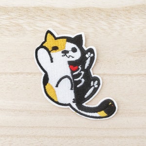 May include: An iron-on patch featuring an embroidered cat with a skeleton on one side and a calico cat on the other. The cat is holding a red heart in its paw.
