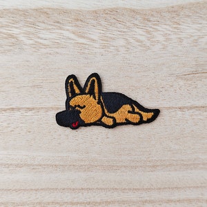Peut inclure: Un patch brodé représentant un berger allemand endormi. Le chien est couché sur le côté, la tête reposant sur ses pattes.