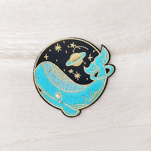 Peut inclure: Écusson brodé représentant une baleine bleue dans un ciel étoilé. La baleine a des détails dorés et est placée sur un cercle noir avec une planète et des étoiles. L'écusson est sur une surface en bois clair.