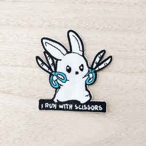 May include: White bunny patch with black outline, holding blue scissors. The patch has the text "I RUN WITH SCISSORS" on a black rectangle. The patch is on a light wood grain surface.