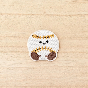 Peut inclure: Un écusson brodé blanc et circulaire représentant un ballon de baseball avec un visage souriant. L'écusson a des détails de couture marron et jaune, et des mitaines marron. L'écusson est sur une surface en bois clair.