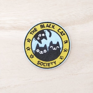 May include: A round, yellow embroidered patch with the text "The Black Cat Society" and three black cats inside a blue circle.