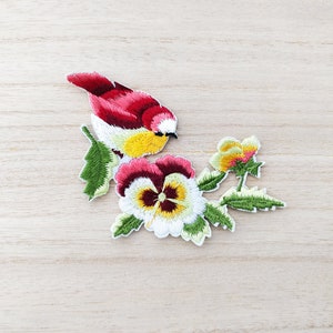 Peut inclure: Patch brodée thermocollante représentant un oiseau rouge et jaune perché sur une branche avec deux pensées blanches et jaunes. Les fleurs ont des centres bordeaux et des feuilles vertes.