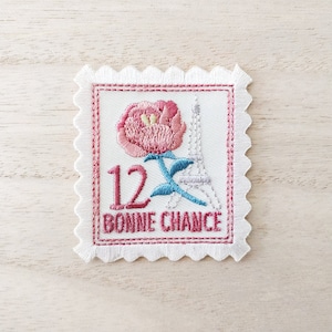 May include: A white embroidered patch with a pink flower, a gray Eiffel Tower, and the text "12 BONNE CHANCE". The patch has a scalloped edge.