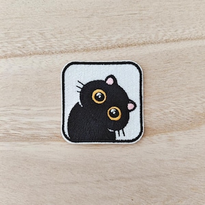 May include: A square embroidered patch featuring a black cat with large, round, yellow eyes and small pink ears. The cat is set against a white background within a thick black border, creating a high-contrast design with detailed stitching.
