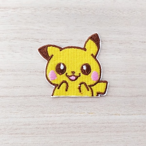 May include: An embroidered patch featuring a cartoon Pikachu, yellow with brown ears and pink cheeks. The patch is displayed on a light brown wooden surface.