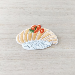 May include: Embroidered patch featuring a sushi design. The patch has a light orange and white color scheme, with a detailed depiction of sushi, including rice, fish, and garnishes. The patch is designed to be ironed on or sewn onto fabric.