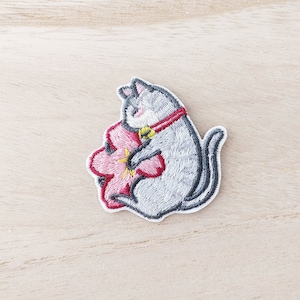 May include: Embroidered patch of a grey cat embracing a pink flower with a yellow centre. The cat wears a red collar with a gold bell. The patch has a white border and is set against a light wood grain background.