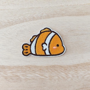 May include: Embroidered patch with a cartoon clownfish design. The patch features an orange body with white stripes, brown outlines, and a small pink blush on the cheek. The patch is approximately 5 cm wide.