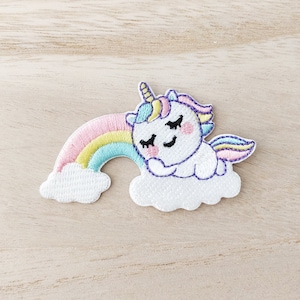 May include: Embroidered patch featuring a sleeping unicorn on a cloud with a rainbow. The unicorn has a white body, a yellow horn, and a pink and purple mane. The rainbow has pink, yellow, and blue stripes. The patch is on a light wood surface.