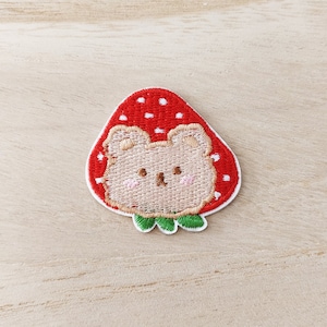 May include: Embroidered iron-on patch featuring a cartoon bear inside a red strawberry with white polka dots. The bear has brown ears, a brown nose, and pink cheeks.