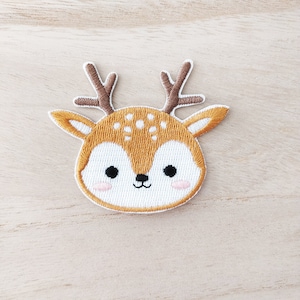 May include: A brown and white embroidered deer head patch with a cute cartoon face. The deer has large eyes, a small nose, and a small mouth. The deer's antlers are brown and white.
