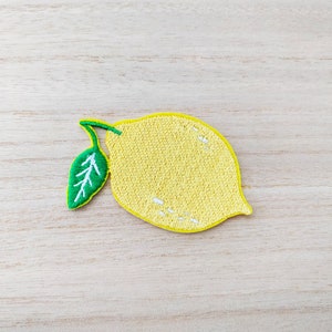 May include: A yellow embroidered lemon with a green leaf, on a light wood background.