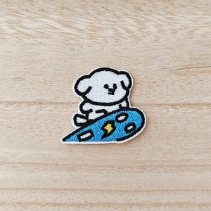 May include: White embroidered patch of a cartoon dog riding a blue surfboard with a yellow lightning bolt design.