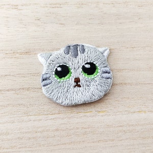 May include: Embroidered iron-on patch of a grey cat with green eyes and a brown nose.
