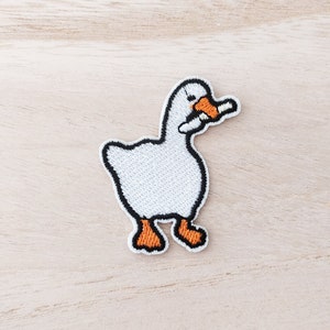 May include: White embroidered goose patch with orange feet and beak holding a white object in its beak.