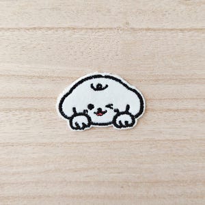 May include: A white embroidered patch of a cartoon dog with a black outline. The dog has a small red mouth, black eyes, and black paws. The patch is on a light brown wooden surface.