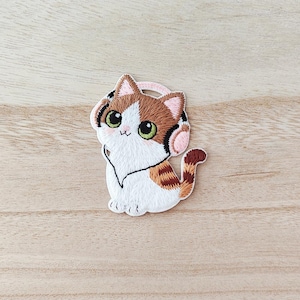 May include: Embroidered patch of a cat wearing headphones. The cat has white fur with brown and orange patches, green eyes, and pink headphones. The patch is designed to be ironed or sewn onto fabric.