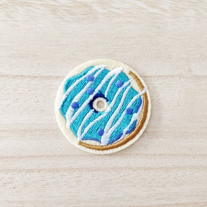 May include: An embroidered donut patch in blue and white with a gold outline. The donut has white icing with blue sprinkles and a hole in the centre.