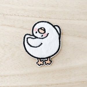 May include: White embroidered iron-on patch of a cartoon bird with black outlines and pink cheeks. The bird is standing with its wings folded and its head tilted to the side.