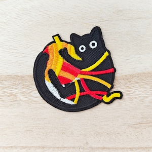 May include: Black cat embroidered patch with yellow and red yarn wrapped around it. The cat has large black eyes.