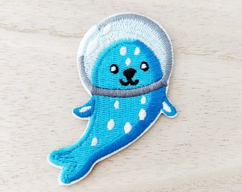 May include: Embroidered patch featuring a cartoon seal in a space helmet. The seal is light blue with white spots and a black nose and mouth. The helmet is gray and white, and the patch has a white border.
