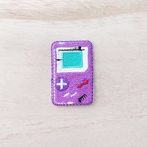 May include: A purple embroidered patch depicting a retro handheld games console. The patch has a teal screen, white detailing, a white plus sign, and red buttons. The rectangular patch has rounded corners and a white border.