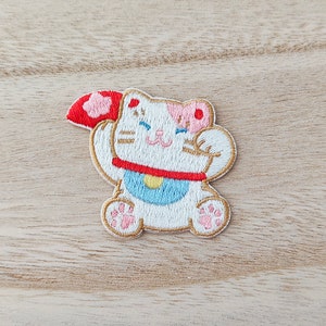 May include: White embroidered patch of a cartoon cat with pink ears and a red and blue collar. The cat is holding up its right paw and has a happy expression on its face.