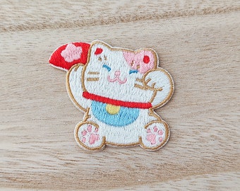 May include: White embroidered patch of a cartoon cat with pink ears and a red and blue collar. The cat is holding up its right paw and has a happy expression on its face.
