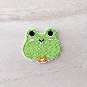 May include: Green embroidered frog patch with black eyes, pink cheeks, and a small orange bow tie.