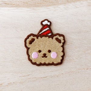 May include: A brown embroidered bear wearing a red and white striped party hat. The bear has pink cheeks and a brown nose.