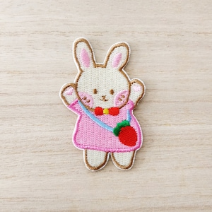 Peut inclure: Patch brodée thermocollante représentant un lapin blanc portant une robe rose, une bandoulière bleue et verte avec une fraise rouge et un nœud papillon rouge.