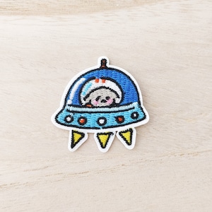 May include: Embroidered patch featuring a blue UFO with a cute alien inside. The UFO has a light blue rim with orange and white accents. The alien is gray with a white hat and pink cheeks. The patch has yellow triangles at the bottom.