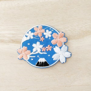 May include: A blue embroidered patch with a white and pink cherry blossom design and a black mountain in the center.