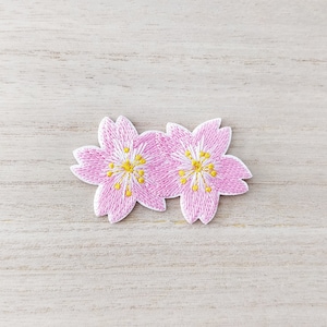 Peut inclure: Écusson brodé représentant deux fleurs de cerisier roses avec des centres jaunes. Les fleurs ont des contours blancs et sont posées sur un fond clair en grain de bois. Idéal pour ajouter aux vêtements ou accessoires.