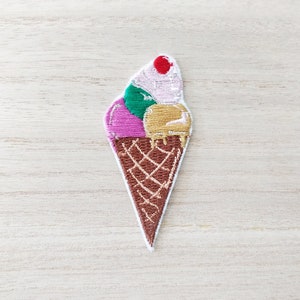 May include: An embroidered iron-on patch of an ice cream cone with three scoops of ice cream. The ice cream is pink, green, and yellow with a red cherry on top.