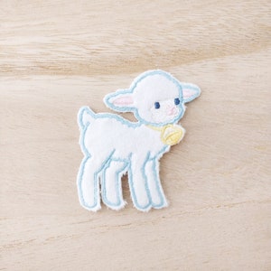May include: Embroidered patch of a white lamb with light blue and pink detailing. The lamb has a yellow bell and is set against a light wood grain background. A decorative patch suitable for clothing or craft projects.
