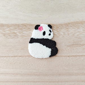 May include: A fluffy panda bear toy with black and white fur, a pink flower on its head, and black eyes. The panda is in a sitting position. The background is a light brown wooden surface.