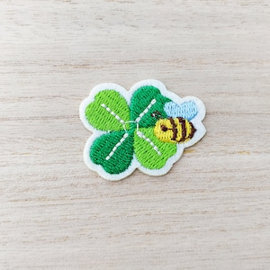 May include: A green four-leaf clover with a bee embroidered on it. The bee is yellow and black with white wings.