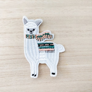 May include: White embroidered alpaca wearing a green and pink scarf. The alpaca is standing on a light wood background.