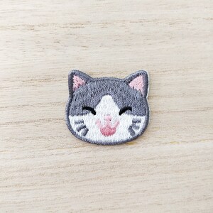 May include: Embroidered patch of a smiling cat's face. The cat features grey fur with white markings, pink inner ears, and a pink tongue. The patch is on a light wood grain surface.