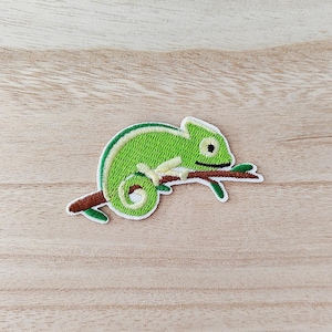 May include: Embroidered patch featuring a green chameleon perched on a brown branch. The chameleon has a white outline and a detailed, textured appearance. The patch is designed for application on clothing or accessories.