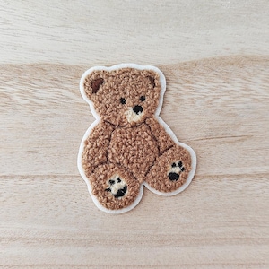 May include: A brown teddy bear patch with a white border. The bear has a textured, fuzzy appearance, black paw prints, and a light brown snout. The patch is likely for clothing or accessories.