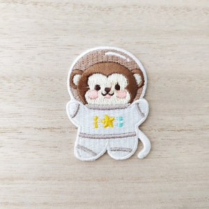 May include: Embroidered iron-on patch of a brown monkey wearing a white astronaut suit with a yellow star on the chest. The monkey has a cute, smiling face.