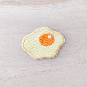 Peut inclure: Un patch brodé jaune en forme d'œuf au plat avec un jaune d'œuf orange et une tache blanche au centre.